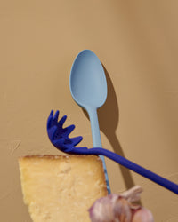 The Slate Spoon Resting on a yellowish background with a Navy Spaghetti spoon in front of it. 